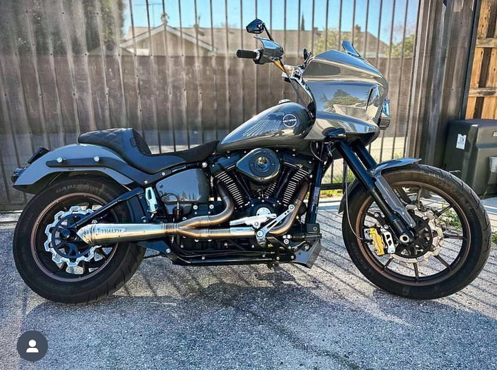 A black motorcycle with the focus on the exhaust system, which is silver in color. The motorcycle is parked against a plain wall backdrop.