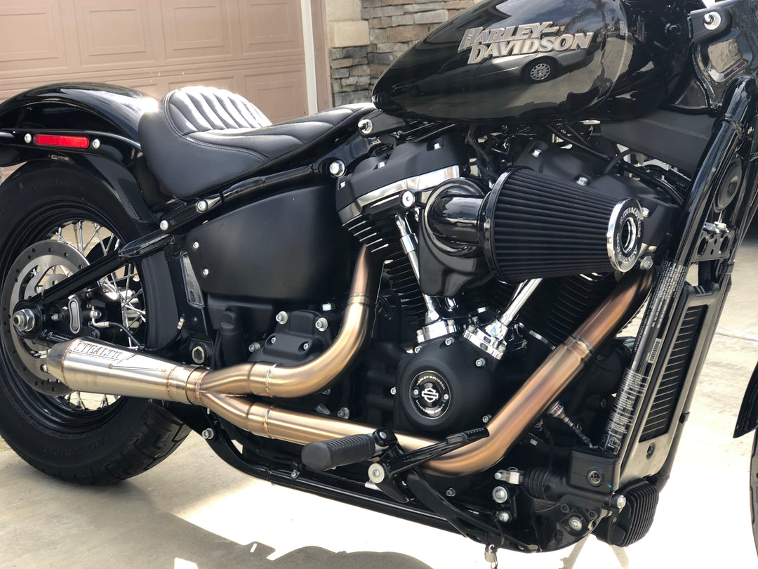 A black motorcycle with the focus on the exhaust system, which is silver in color. The motorcycle is parked against a plain wall backdrop.