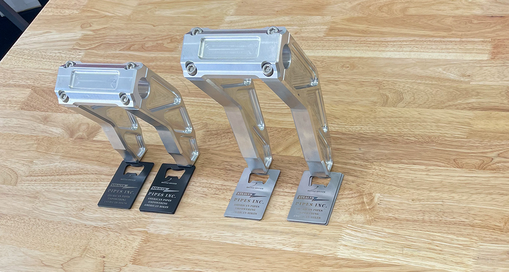 A pair of silver motorcycle bar risers placed on a wooden surface, with engraved details and ratios on the side.