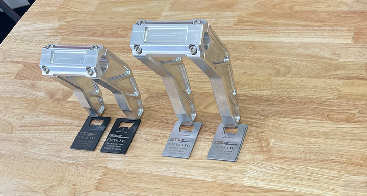 A pair of silver motorcycle bar risers placed on a wooden surface, with engraved details and ratios on the side.