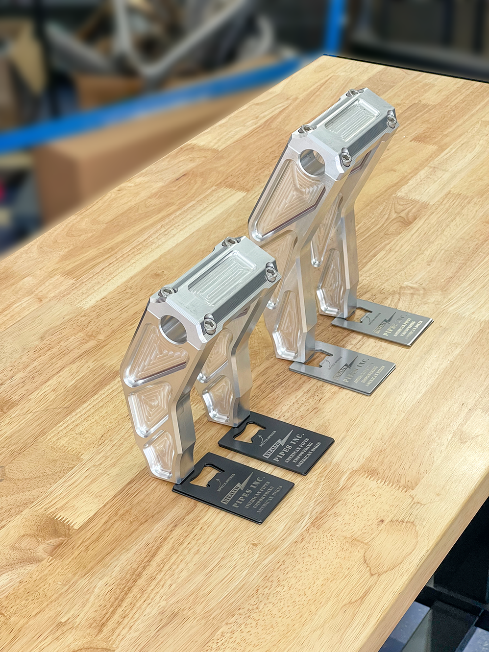 A pair of silver motorcycle bar risers placed on a wooden surface, with engraved details and ratios on the side.