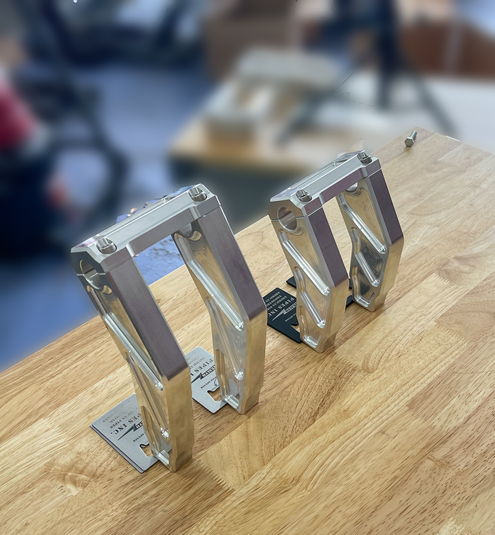 A pair of silver motorcycle bar risers placed on a wooden surface, with engraved details and ratios on the side.