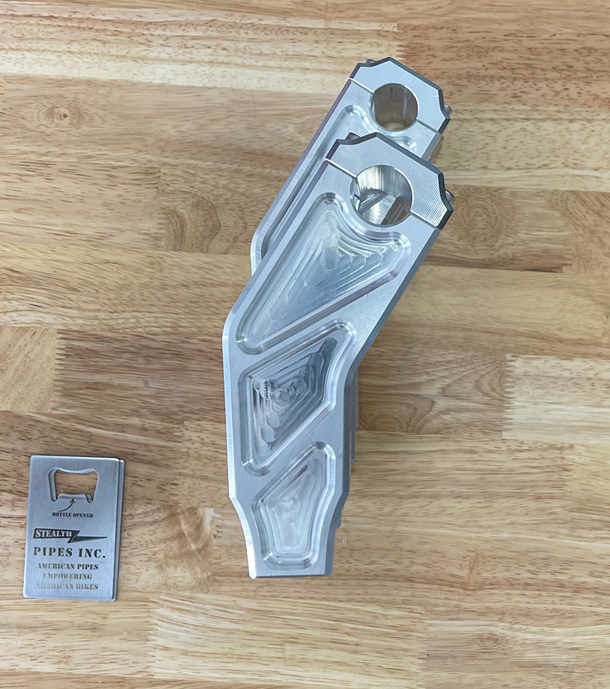 A pair of silver motorcycle bar risers placed on a wooden surface, with engraved details and ratios on the side.