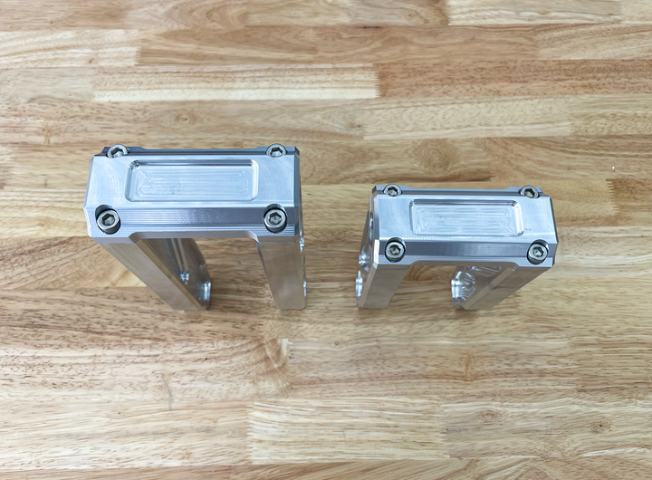 A pair of silver motorcycle bar risers placed on a wooden surface, with engraved details and ratios on the side.