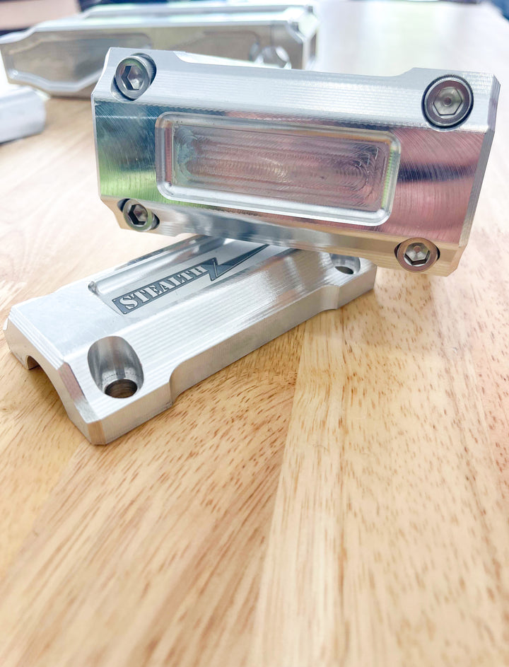 A pair of silver motorcycle bar risers placed on a wooden surface, with engraved details and ratios on the side.